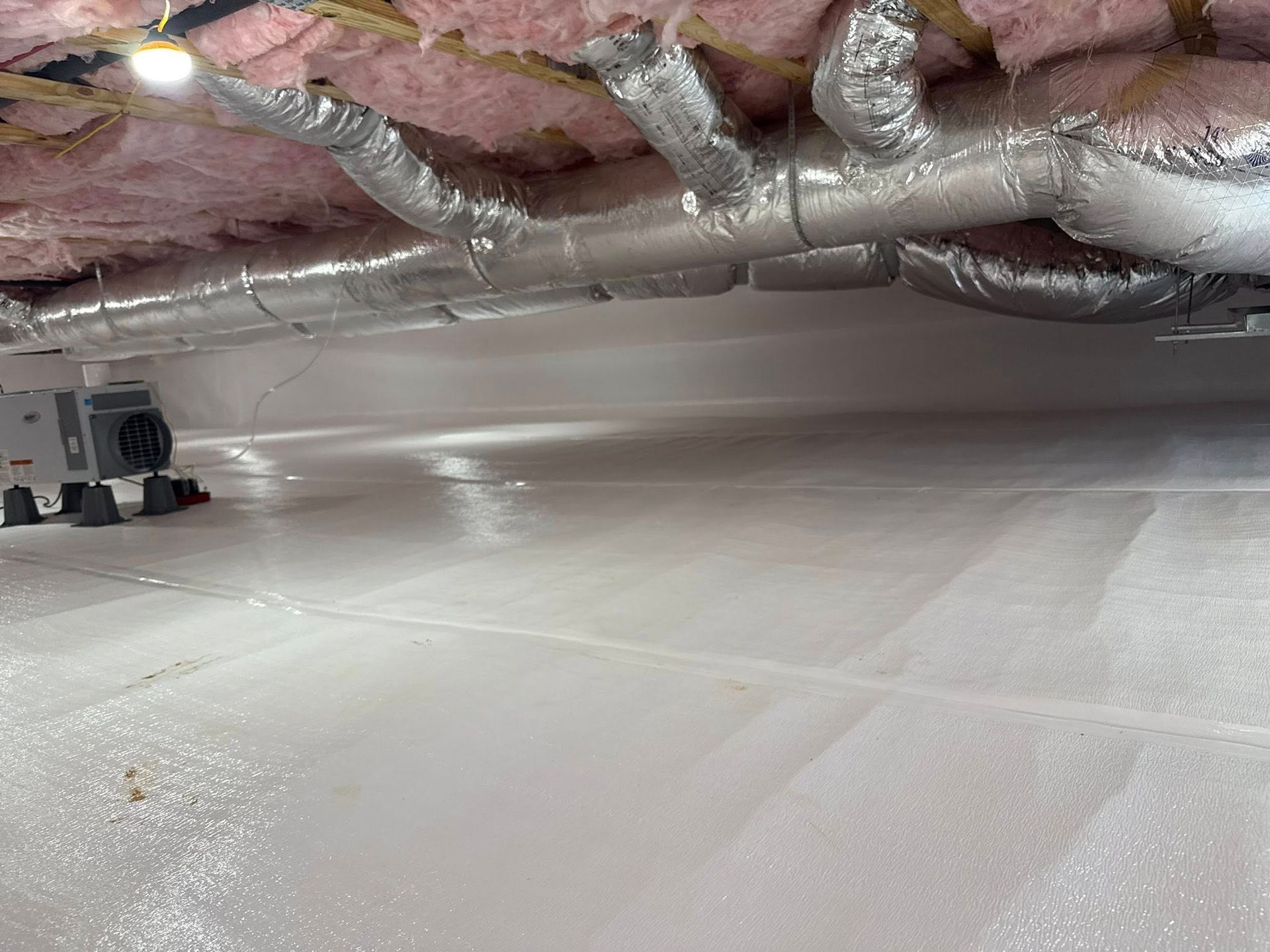 Crawl space with a white vapor barrier floor, pink fiberglass insulation, insulated HVAC ducting, and a dehumidifier.