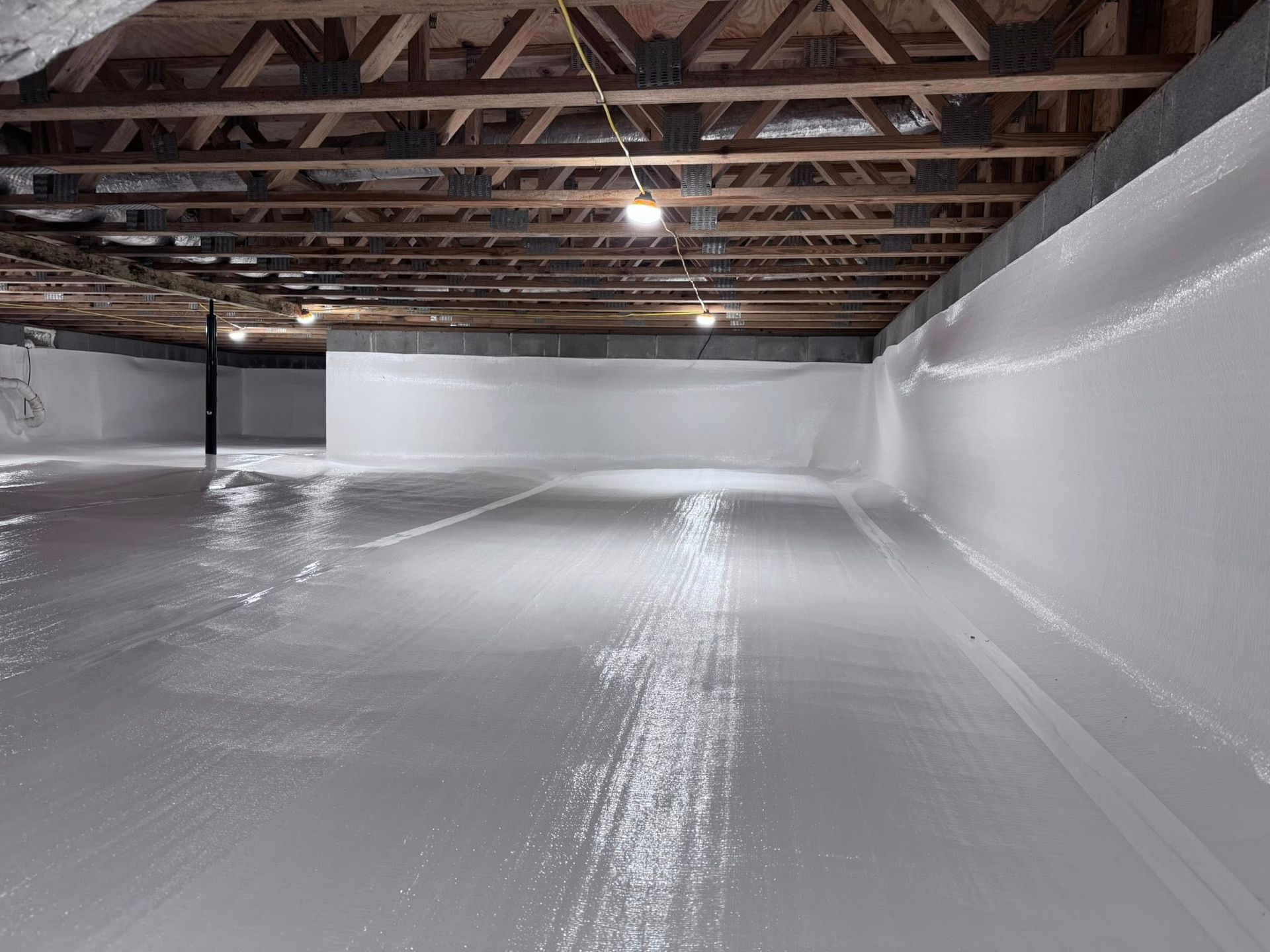 A crawl space with white moisture-barrier sheeting installed on the floor and walls beneath exposed wooden floor joists.