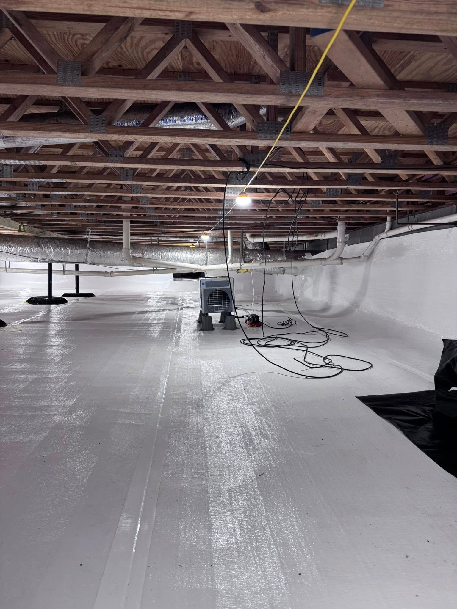 A large attic space with wooden trusses above and a white, smooth-coated floor, featuring a dehumidifier in the center.
