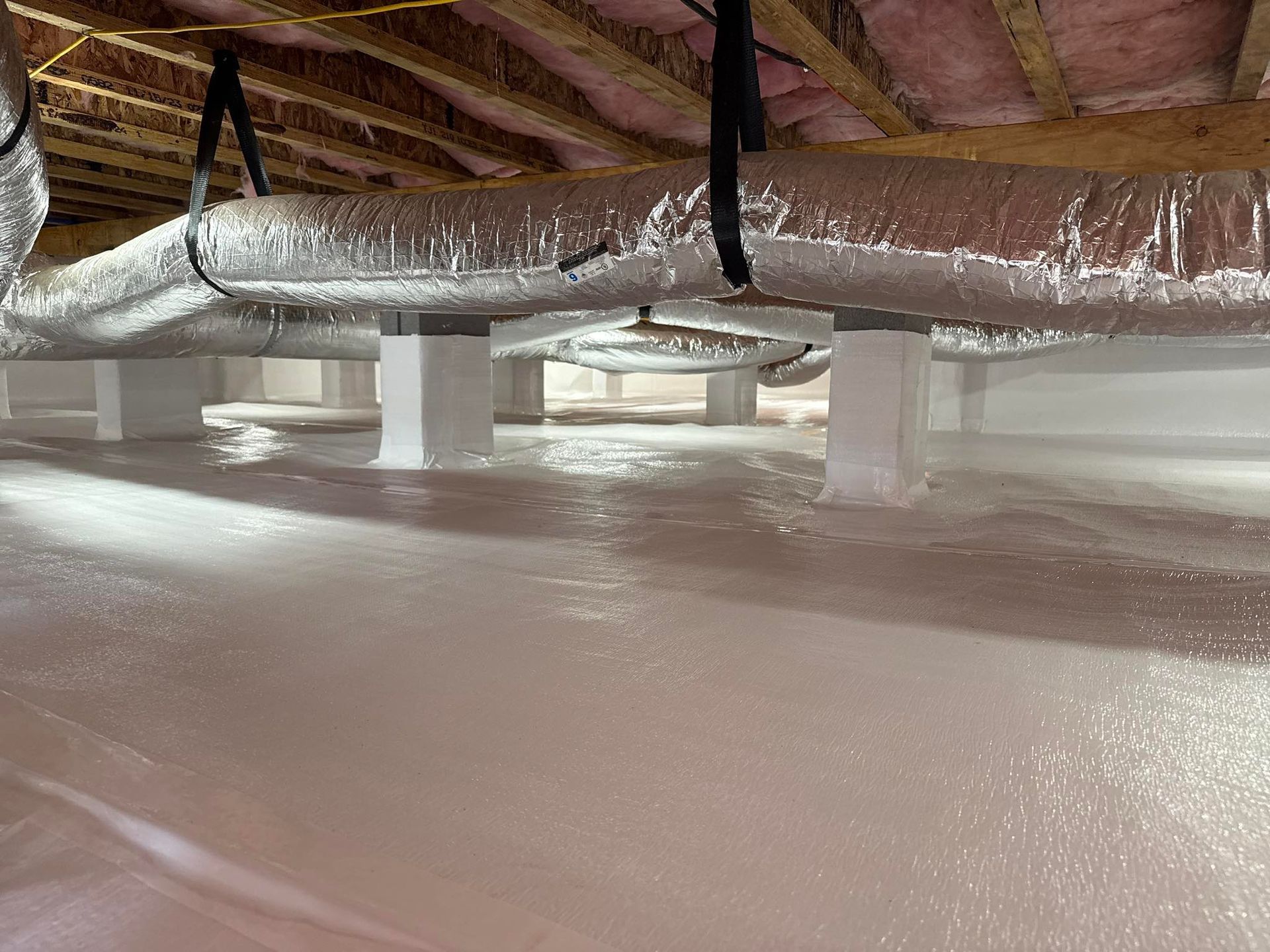 A clean crawl space with a white vapor barrier covering the floor and white-wrapped support pillars under insulated ducts.