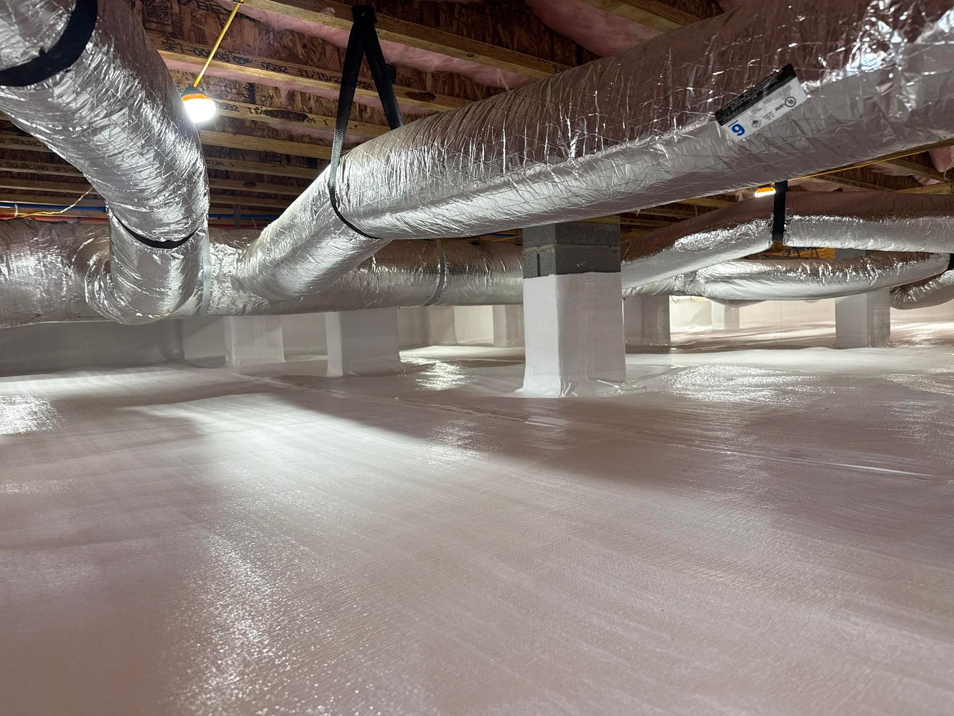A crawl space with insulated air ducts hanging from ceiling joists over a white vapor barrier covering the ground.