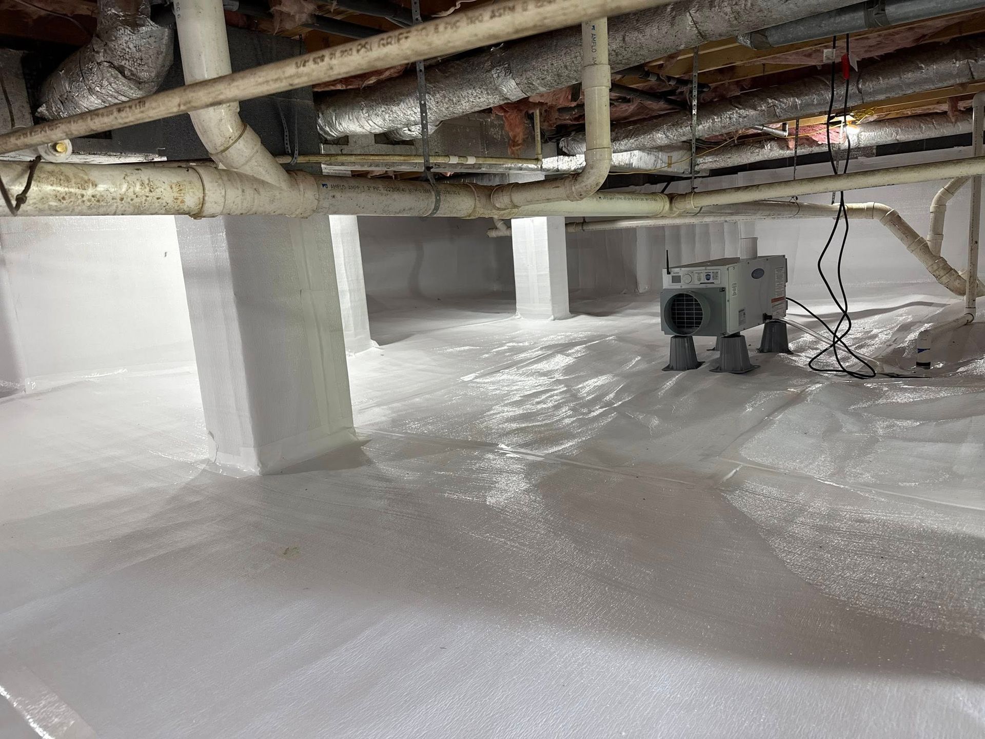 A crawl space encapsulated with white vapor barrier, featuring support columns, exposed plumbing, and a dehumidifier.