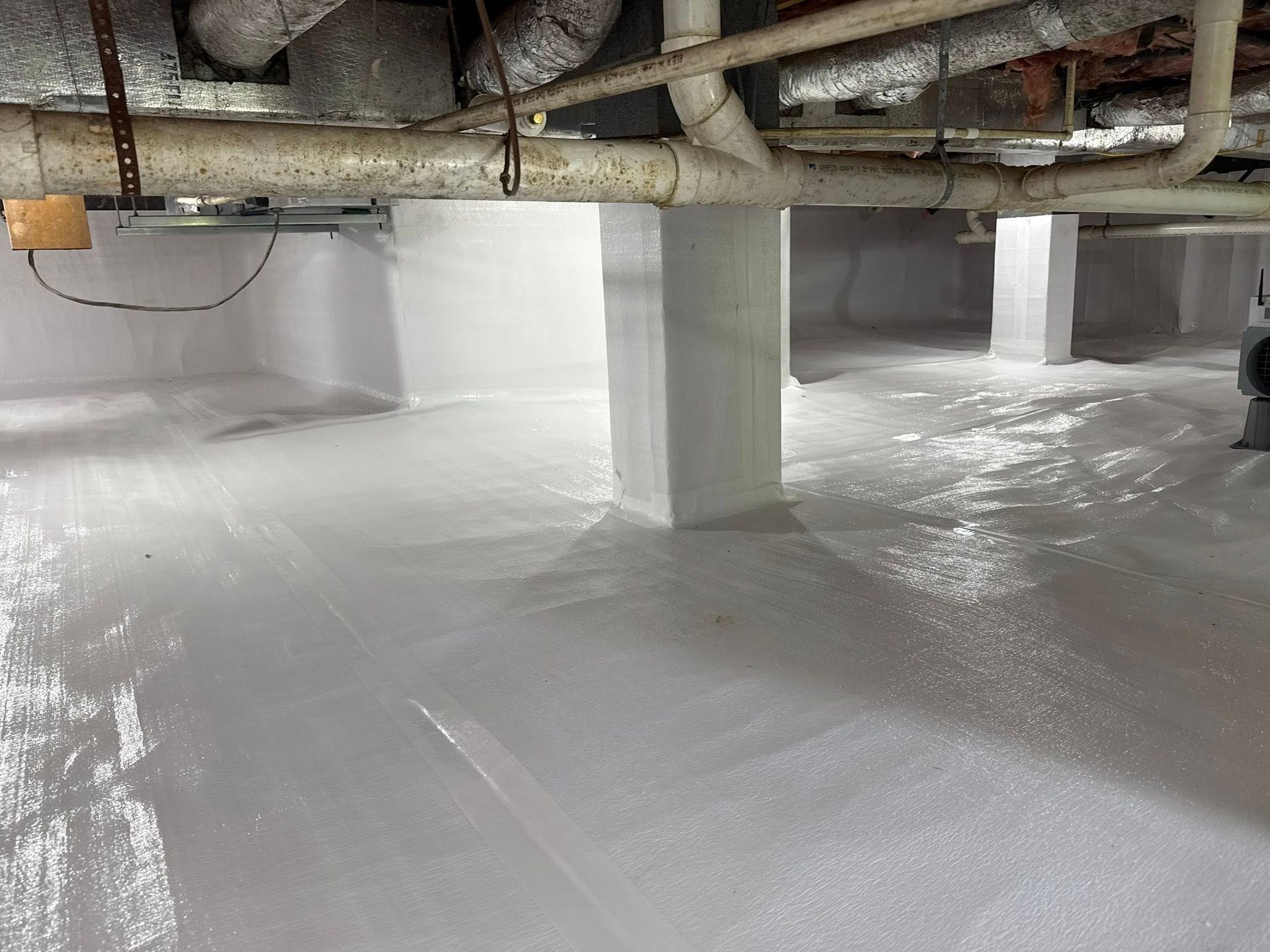 A clean crawl space with white vapor barrier covering the floor and foundation walls, featuring support pillars and pipes.