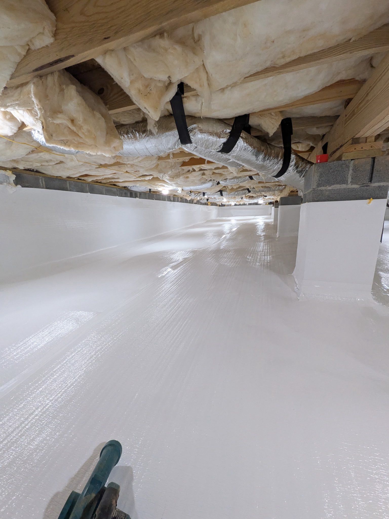 A crawl space with a freshly white-coated floor and foundation walls, featuring exposed floor joists and insulation above.