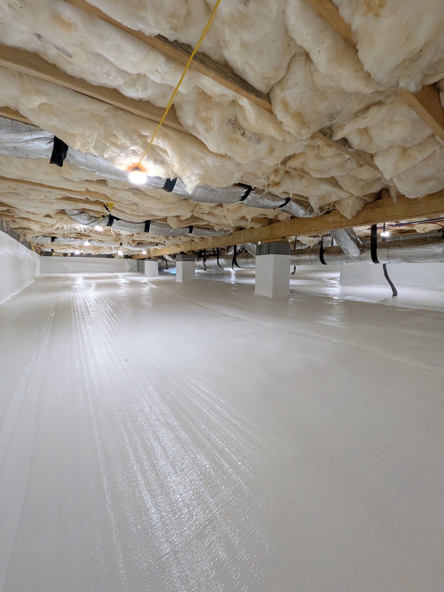 Crawl space with a bright white vapor barrier on the floor and fiberglass insulation between overhead wooden joists.