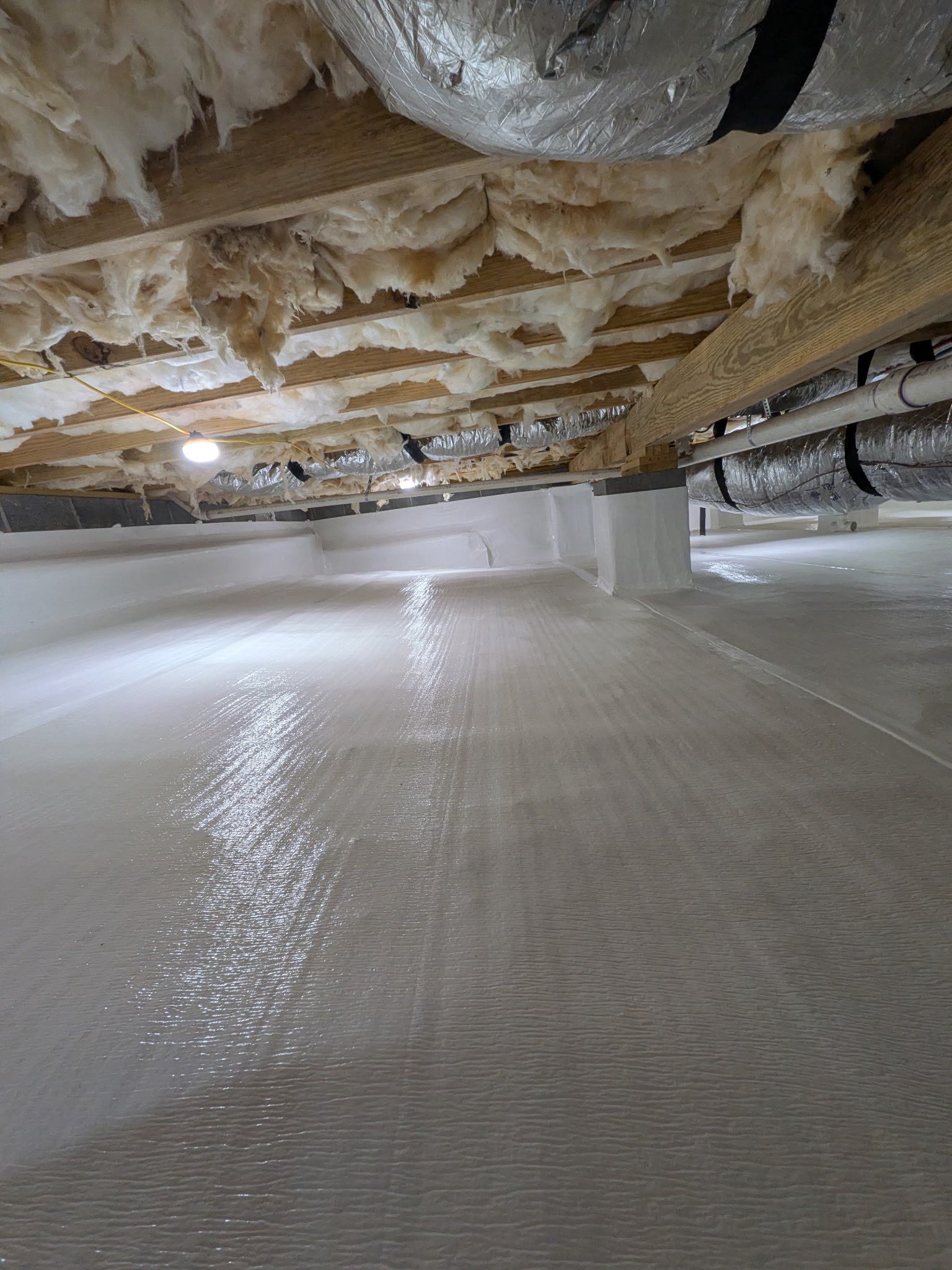 A crawl space with a clean, white vapor barrier on the floor and insulation between exposed wooden ceiling joists.