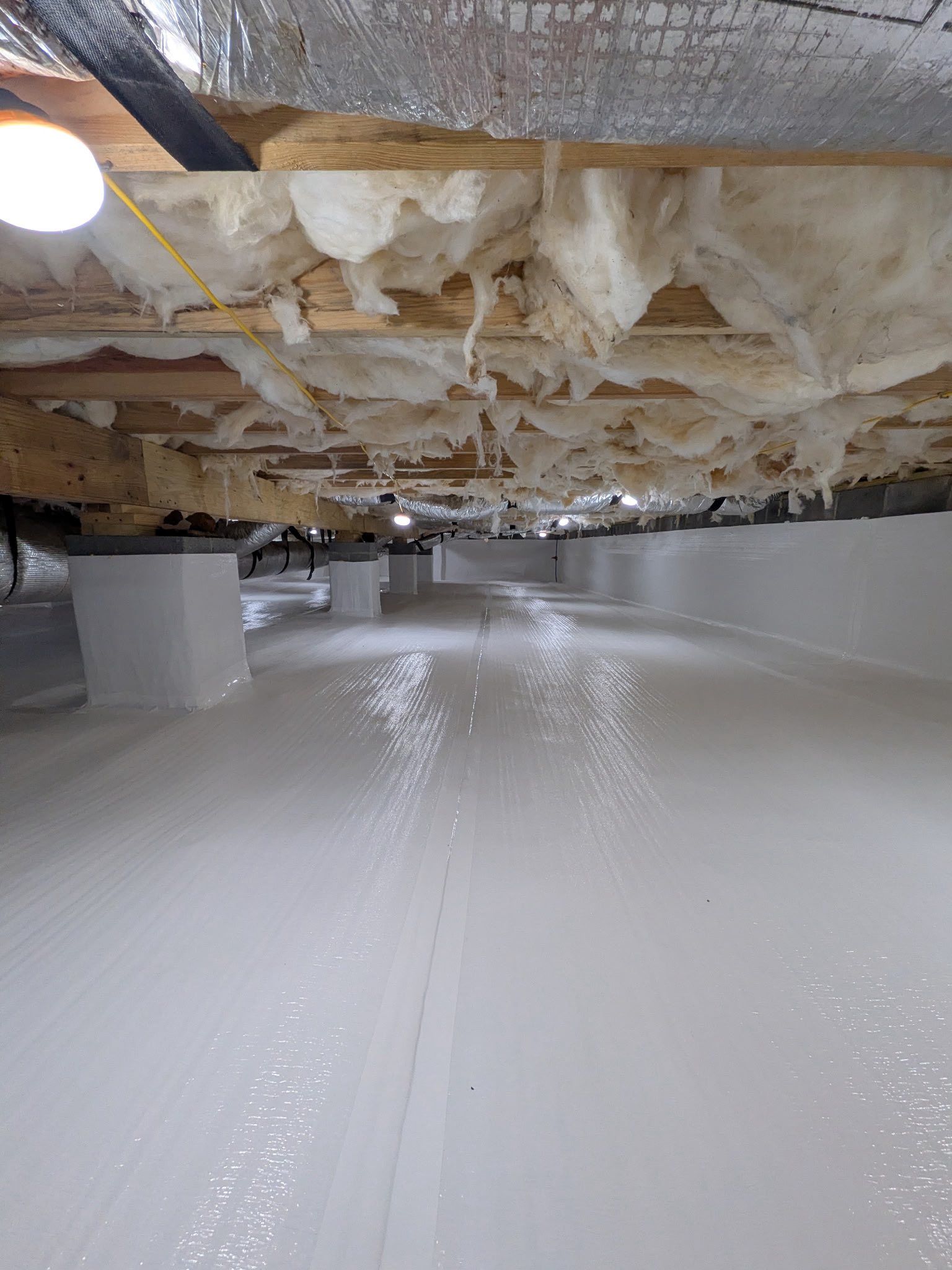 A crawl space with white moisture-barrier flooring, white foundation piers, and insulation installed between floor joists.
