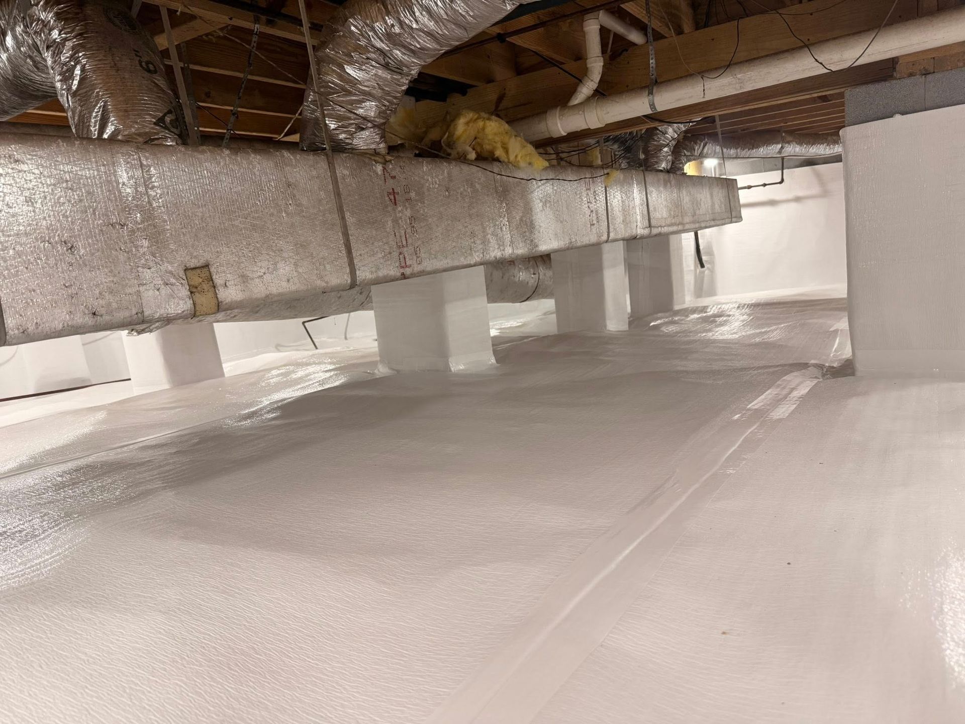 A crawl space with white-coated concrete foundations, a vapor barrier on the floor, and overhead ductwork.