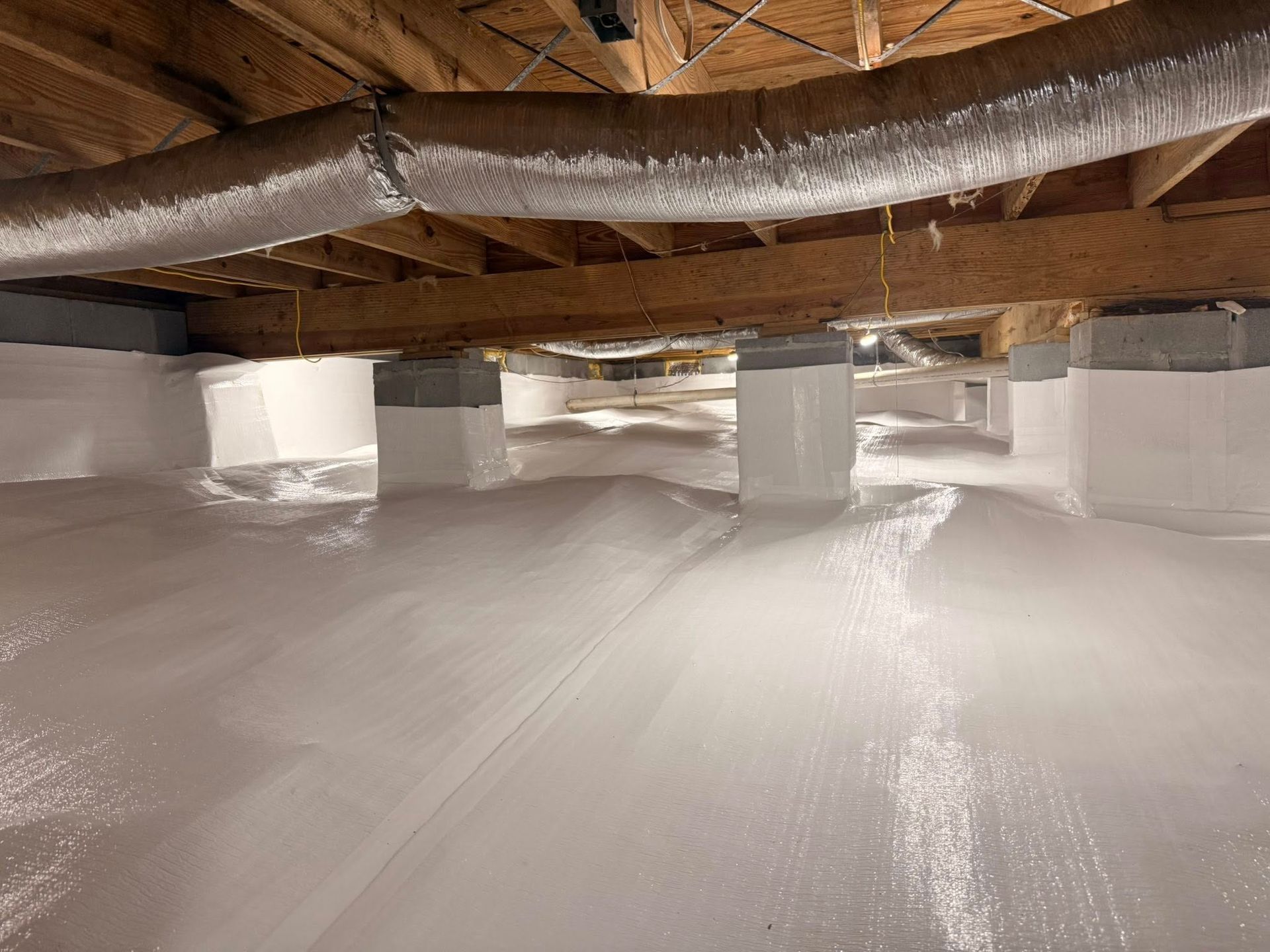 A crawl space encapsulated with white vapor barrier plastic, showing floor, support pillars, and overhead wood joists.