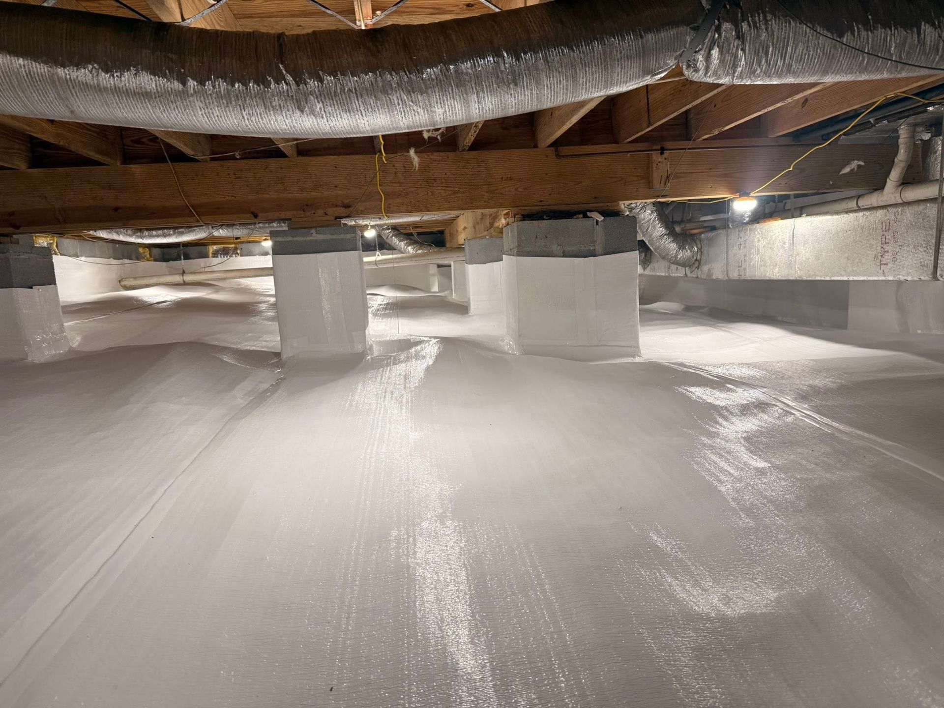 A crawl space encapsulated with white vapor barrier plastic wrapping the floor and support pillars under wooden beams.