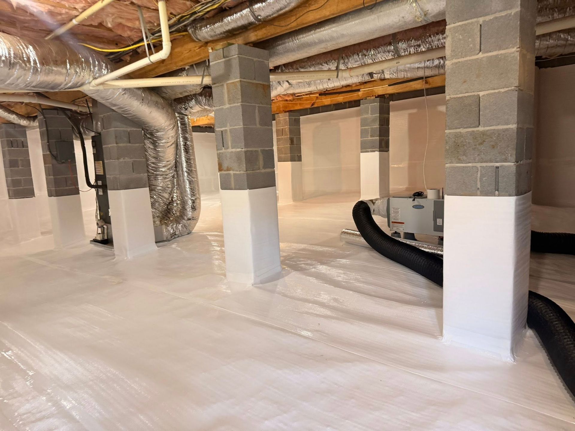 A crawl space with white vapor barriers, block support columns, HVAC ducts, and a dehumidifier system.