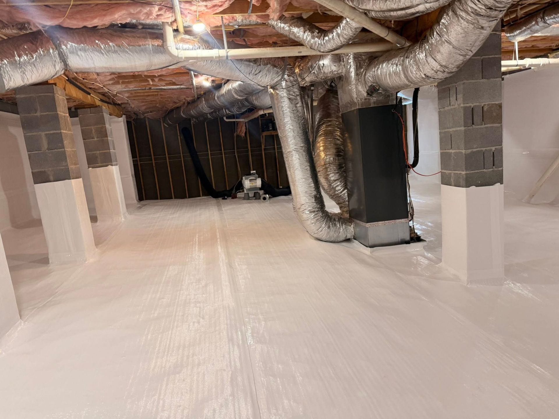 Crawl space with white vapor barrier flooring and walls covering support pillars, under ducts and exposed joists.