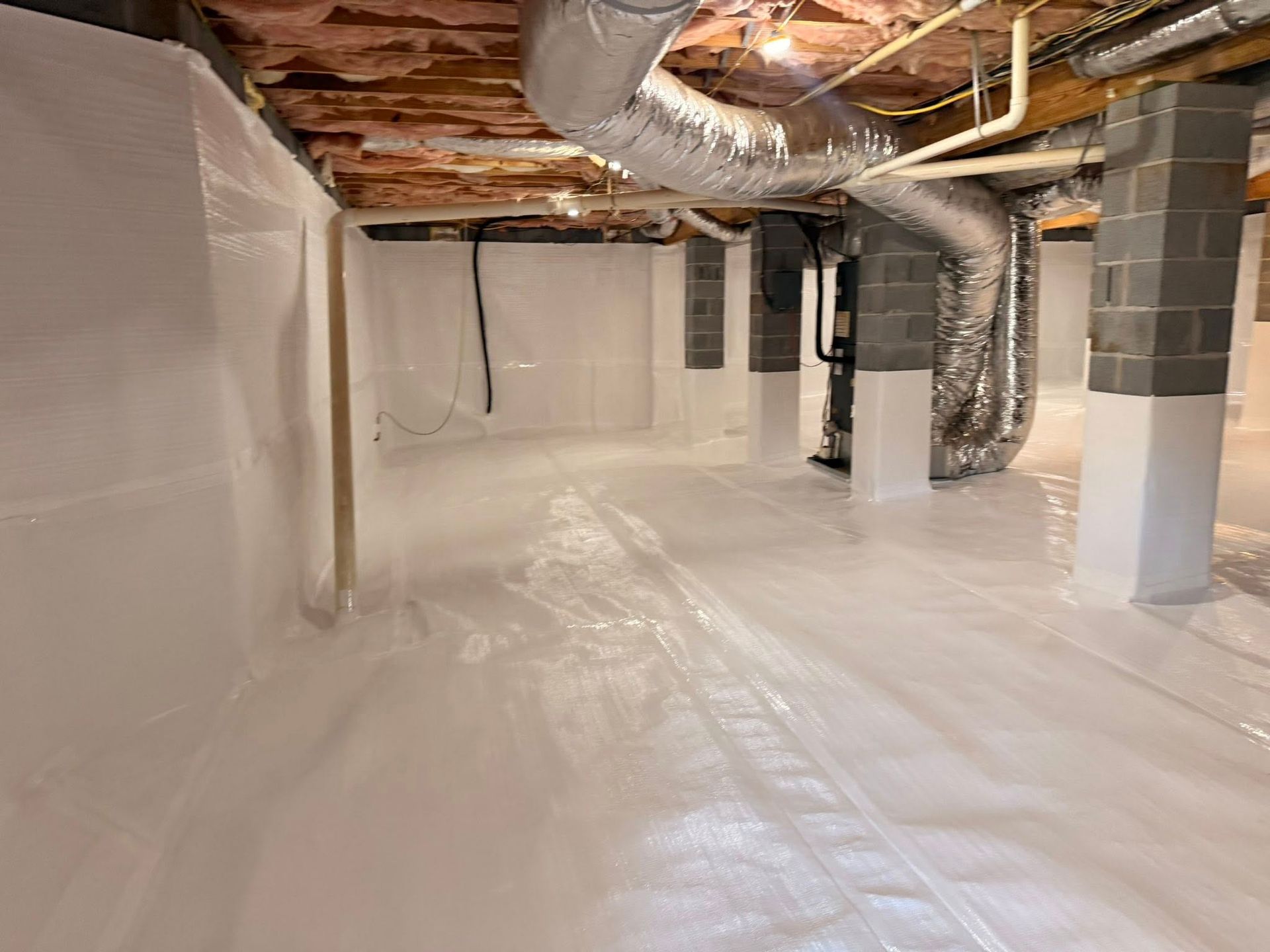 A crawl space with white plastic vapor barrier covering the walls and floor, featuring support pillars and ductwork.