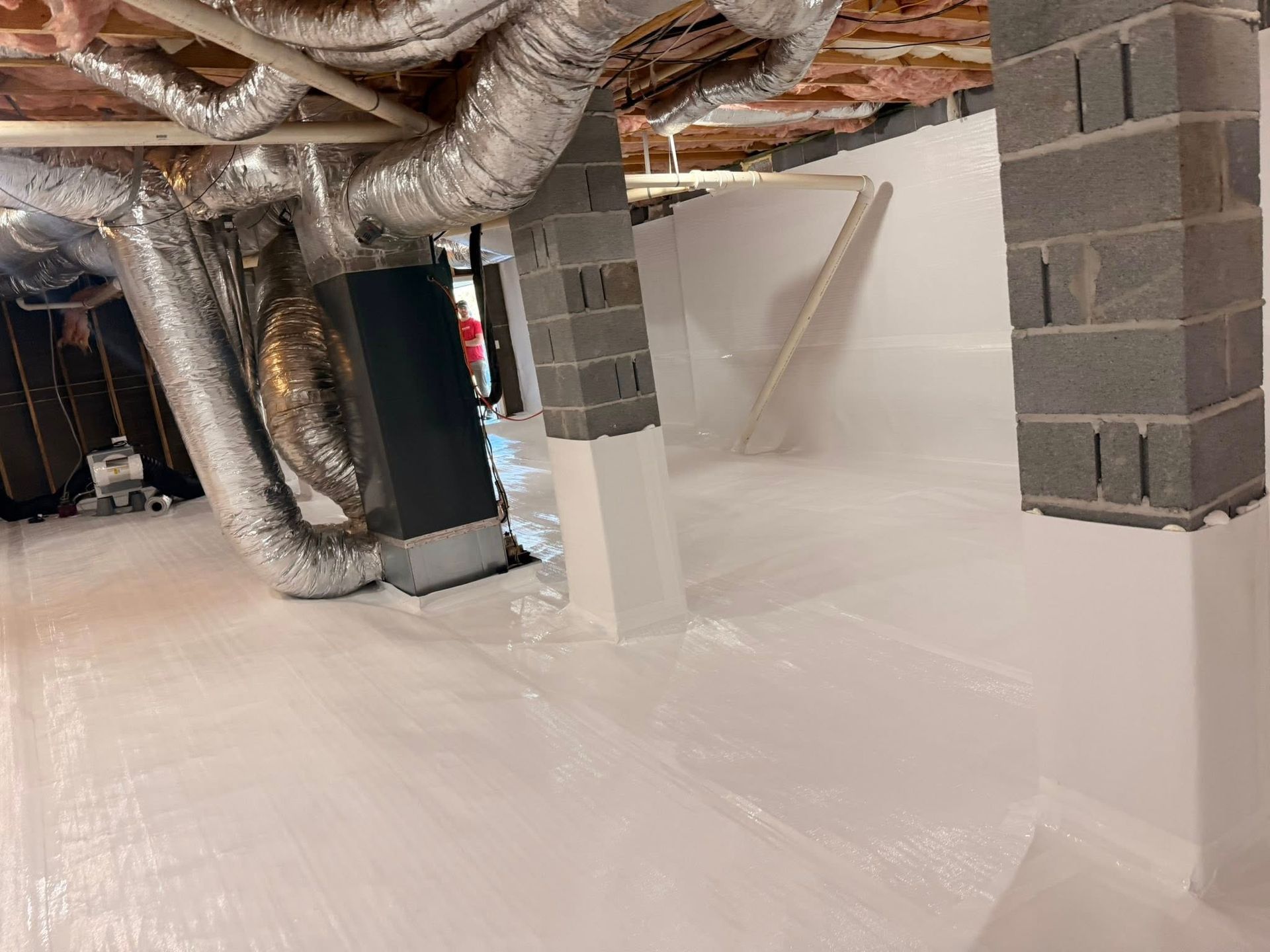 A crawl space featuring a white vapor barrier covering the ground, wrapped support columns, and overhead metallic ducts.