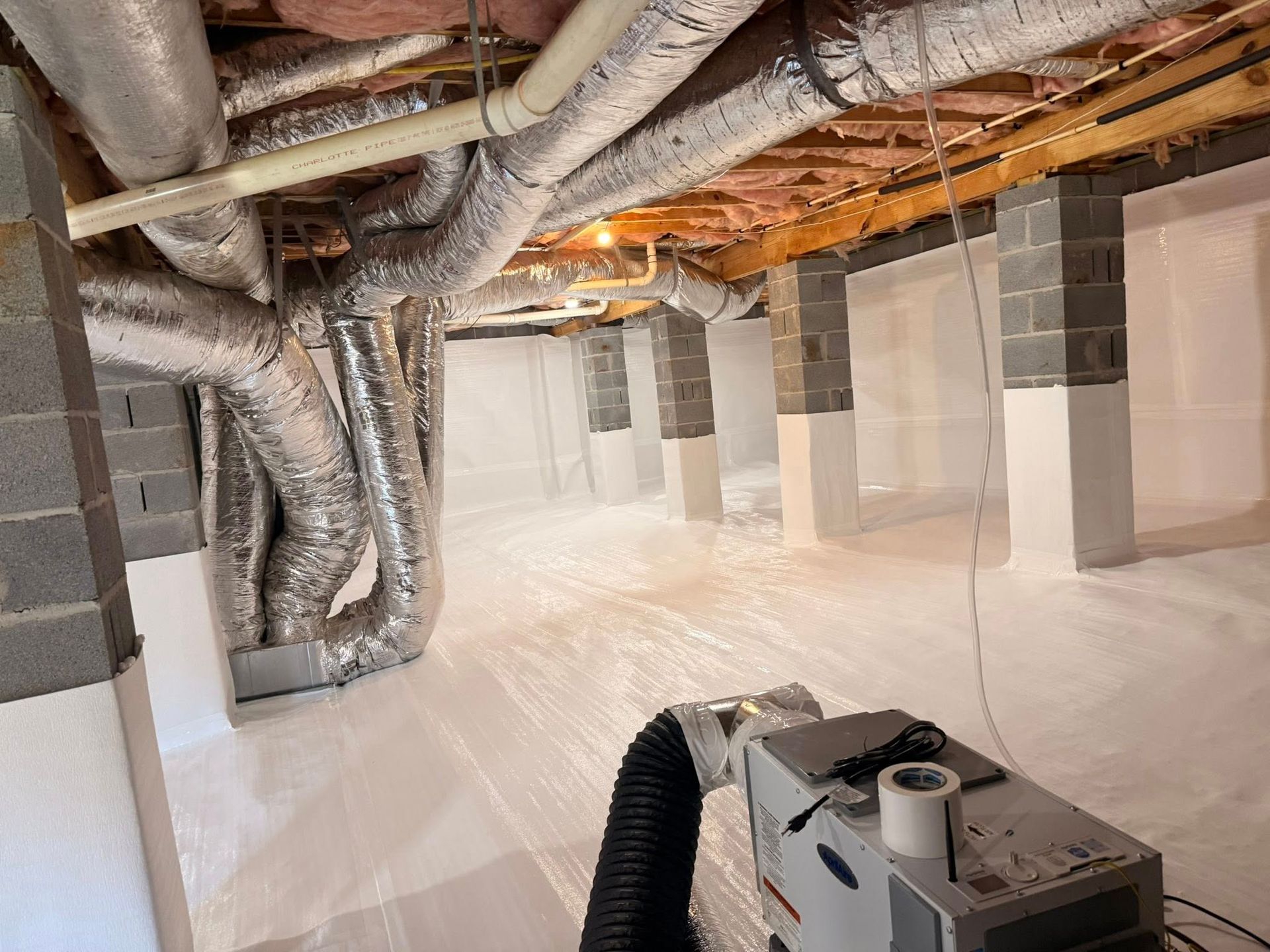 Crawl space with a white moisture barrier on floor and walls, insulated metal ductwork overhead, and a dehumidifier.