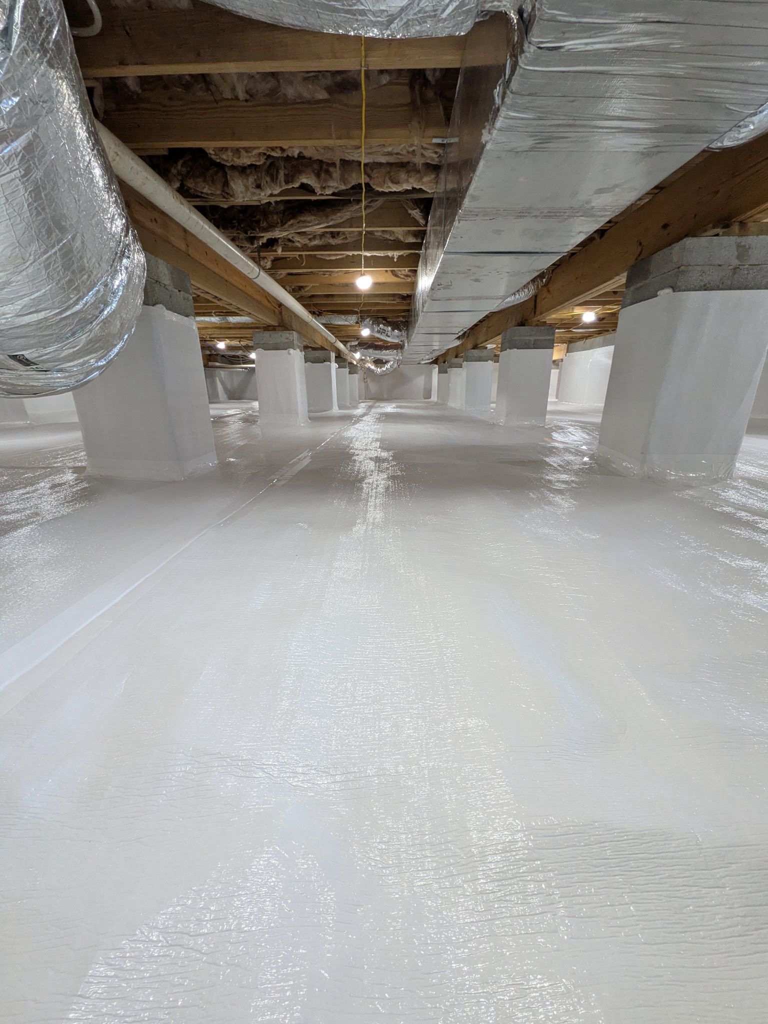 Crawl space with a white vapor barrier covering the floor and wrapping around support piers beneath exposed floor joists.