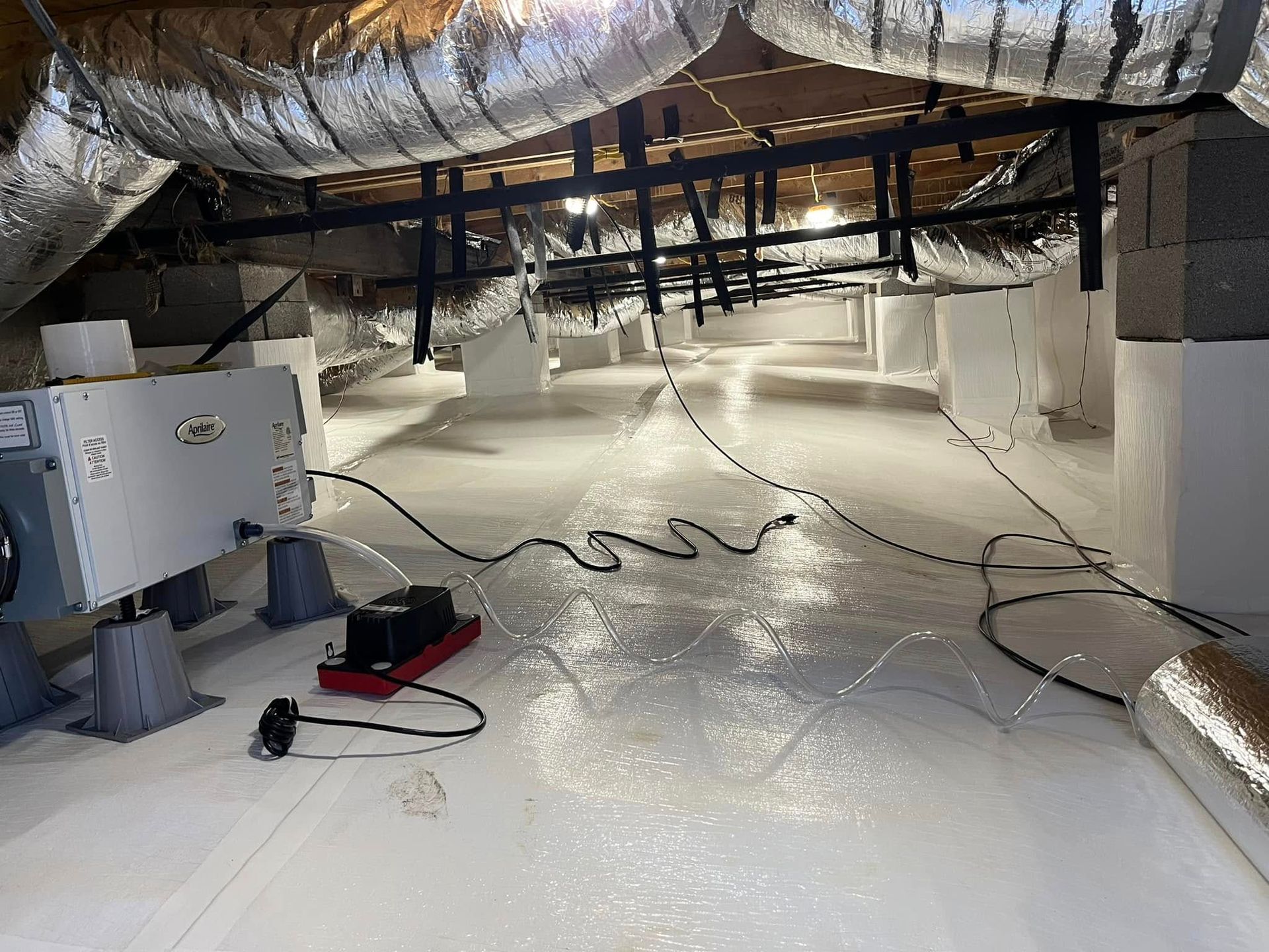 A crawl space with white vapor barrier flooring, a dehumidifier, and silver HVAC ducts hanging from the ceiling joists.