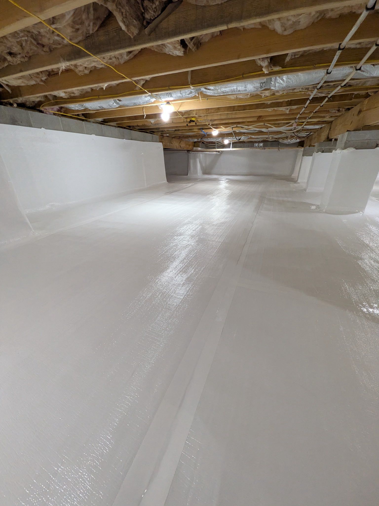 A crawl space with white vapor barrier flooring and walls, and visible wooden floor joists and insulation above.