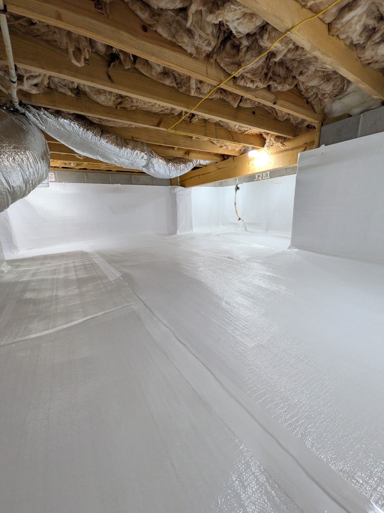 A crawl space with insulated wooden ceiling joists and walls and floors covered in white vapor barrier sheeting.