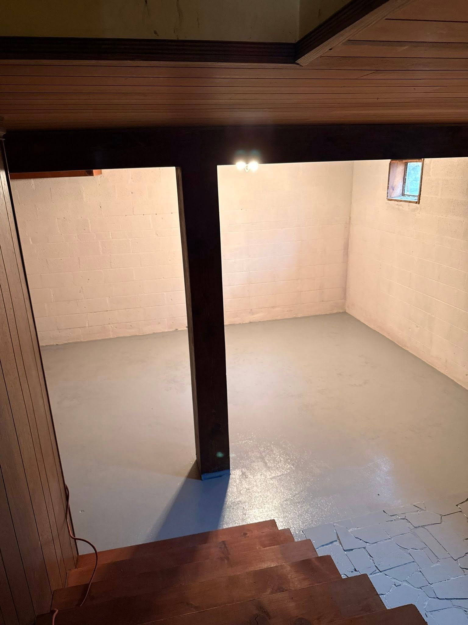 A view looking down wooden stairs into a basement with painted concrete floors, white brick walls, and a central pillar.