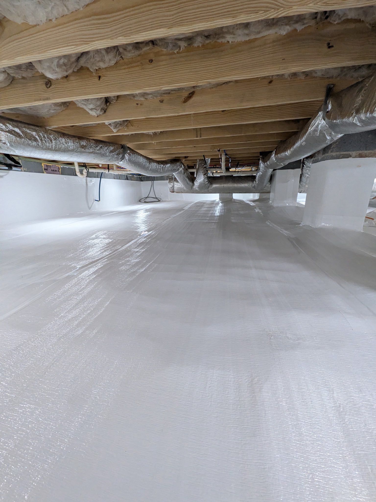 A crawl space with white moisture-barrier flooring, silver ductwork, and exposed wooden floor joists above.