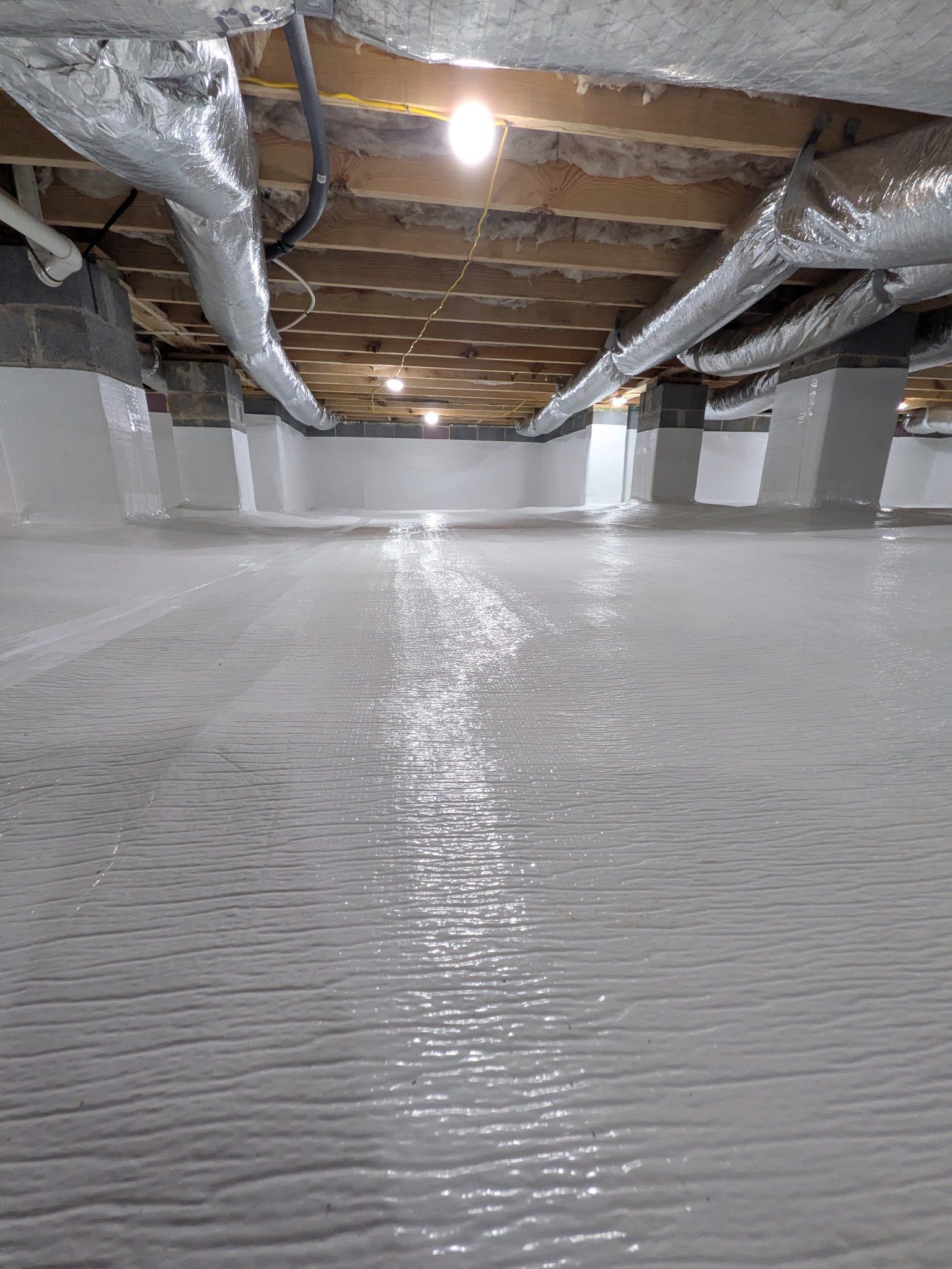A encapsulated crawl space with white vapor barrier flooring and walls, silver insulated ductwork, and wooden joists.