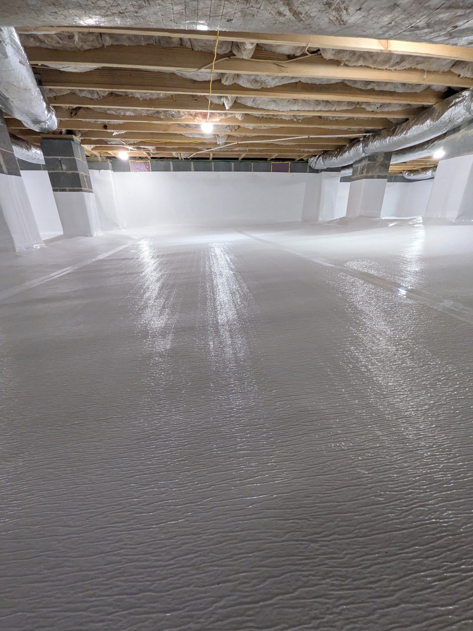 A basement with a freshly applied, reflective gray epoxy floor, white walls, and an exposed wooden ceiling with insulation.