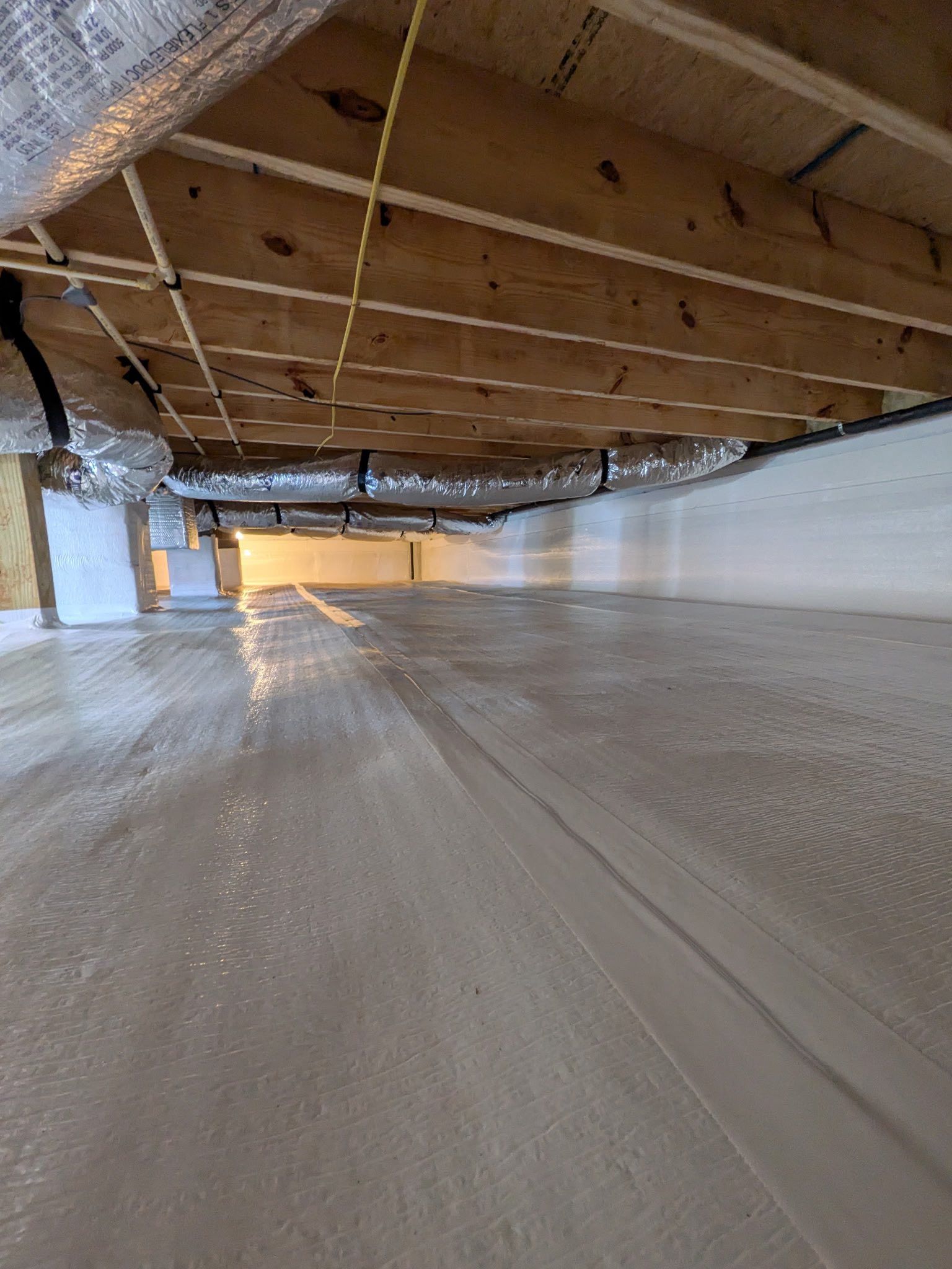 A crawl space with a clean, light-colored vapor barrier on the ground, wooden floor joists, and insulated ducts above.