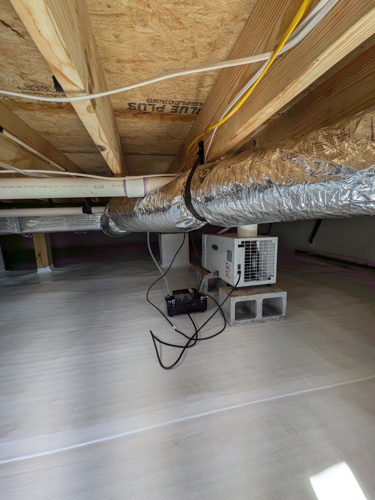 Dehumidifier and drainage pipe located in a clean, encapsulated crawl space with exposed joists above.