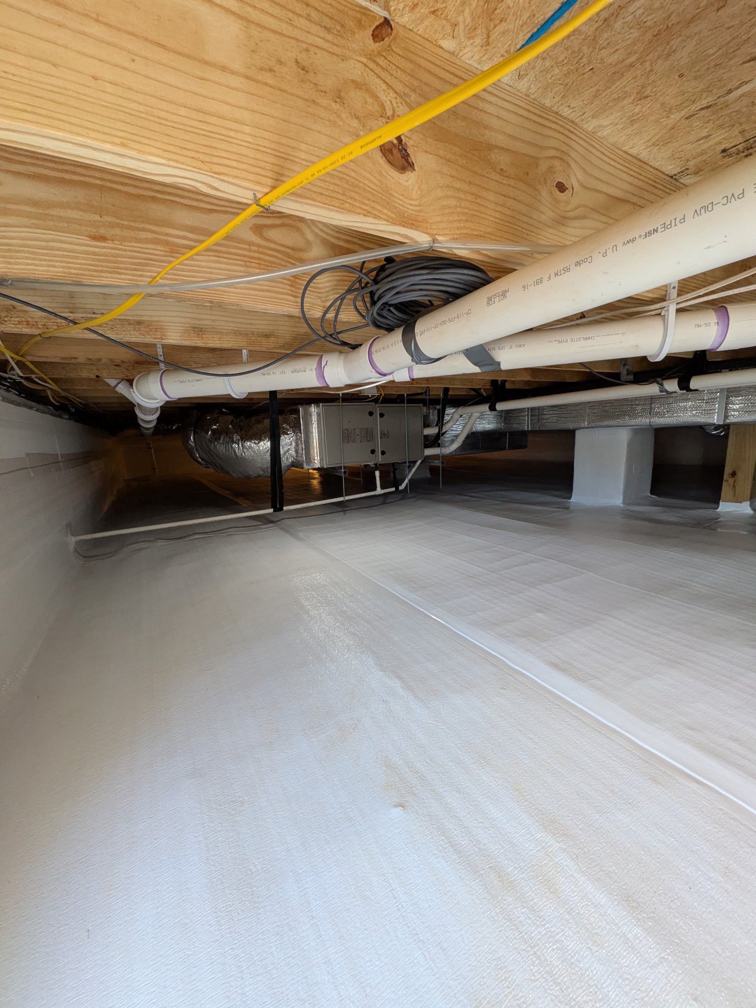A view inside a crawl space with a clean white vapor barrier floor, plumbing pipes, ductwork, and exposed floor joists.