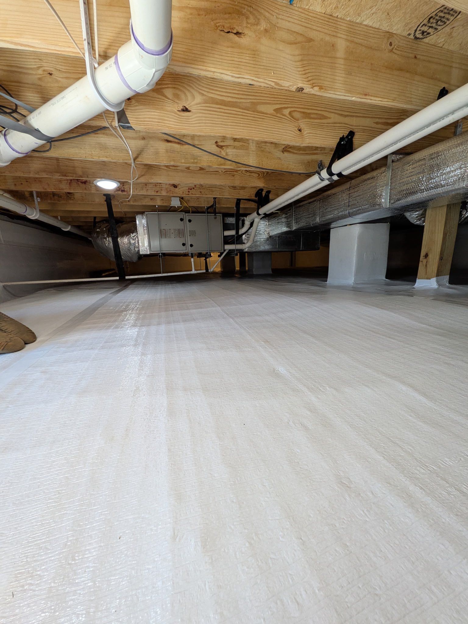 A crawl space featuring a white vapor barrier on the floor, wooden floor joists above, white plumbing pipes, and ductwork.