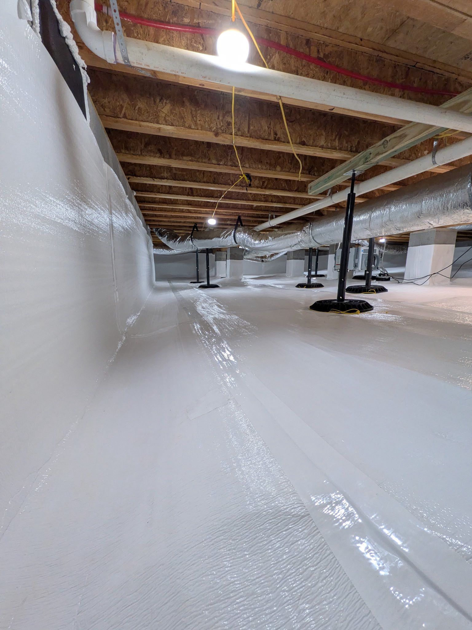A crawl space encapsulated with white vapor barrier plastic on the floor and walls, with overhead joists and ductwork.