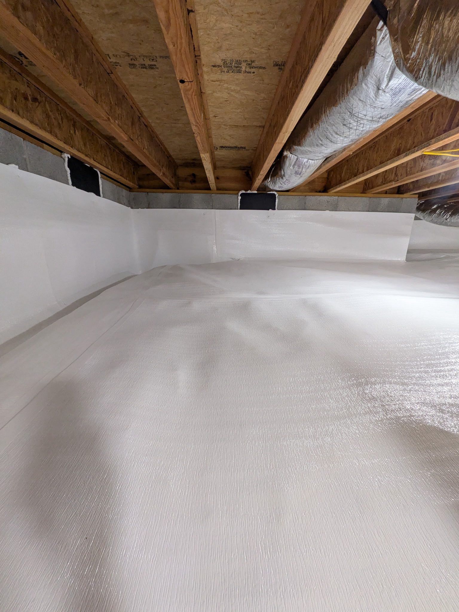 A crawl space with a clean white vapor barrier covering the floor and walls, below wooden floor joists and insulation.