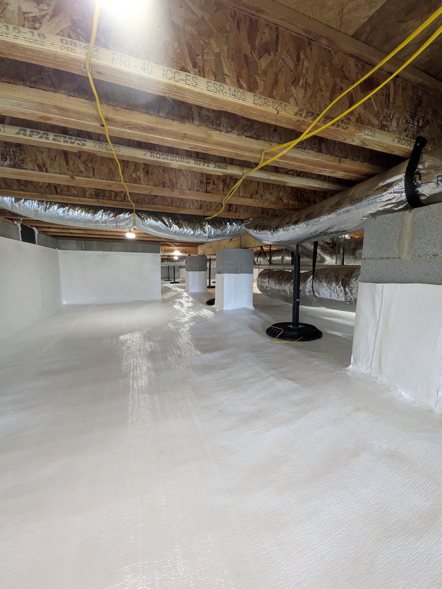 A crawl space with a white vapor barrier on the floor and walls, exposed floor joists, and insulated silver ductwork above.