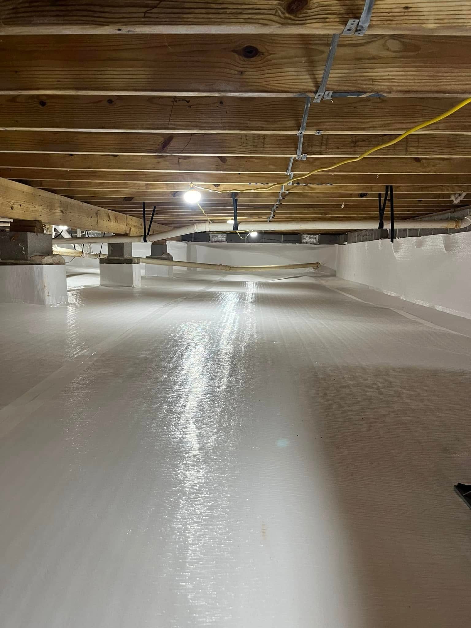 A crawl space with white vapor barrier flooring under exposed wooden ceiling joists, illuminated by a single bright light.