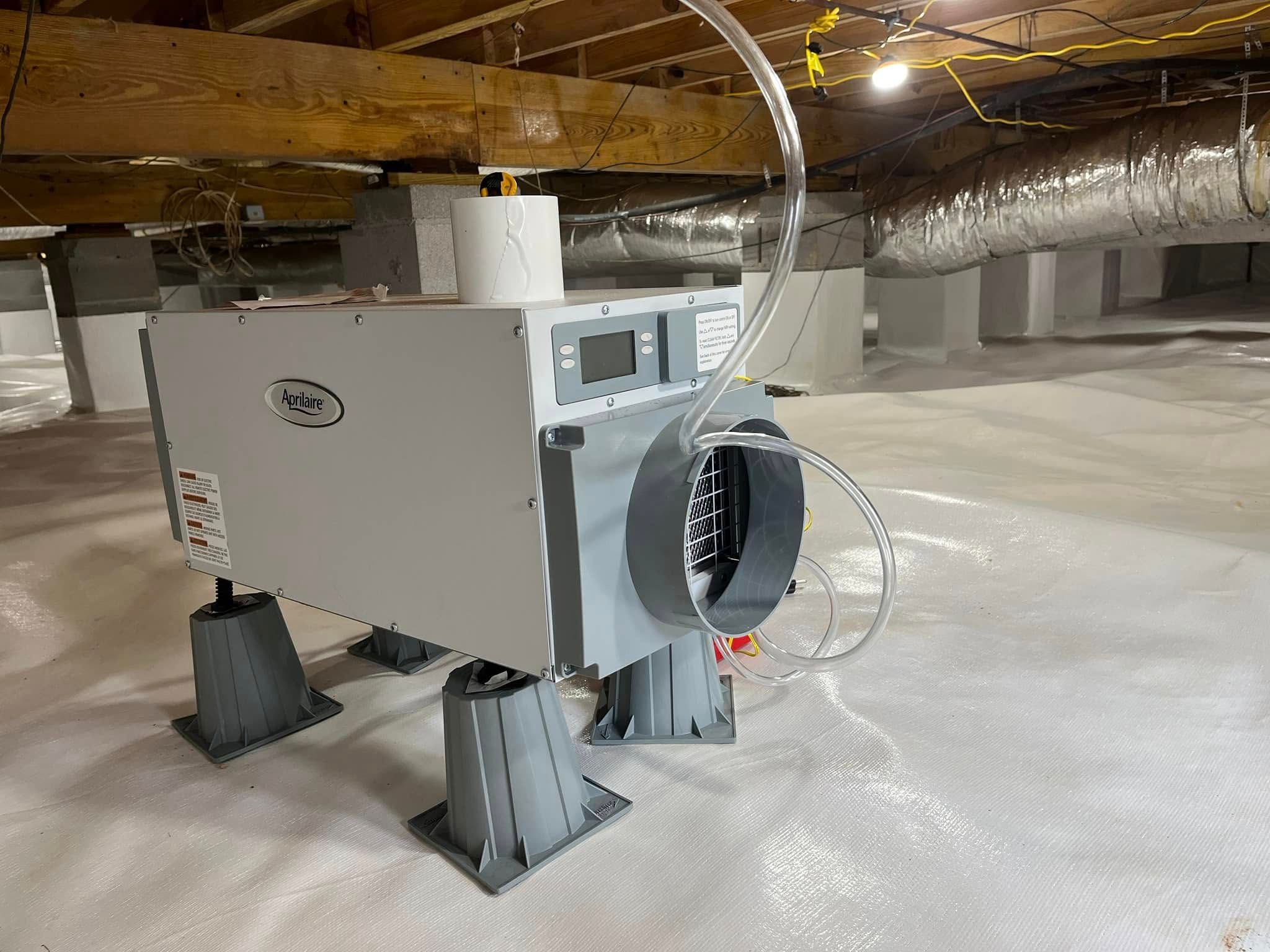 A dehumidifier on gray plastic stands inside a crawl space with a white vapor barrier and exposed wooden joists.