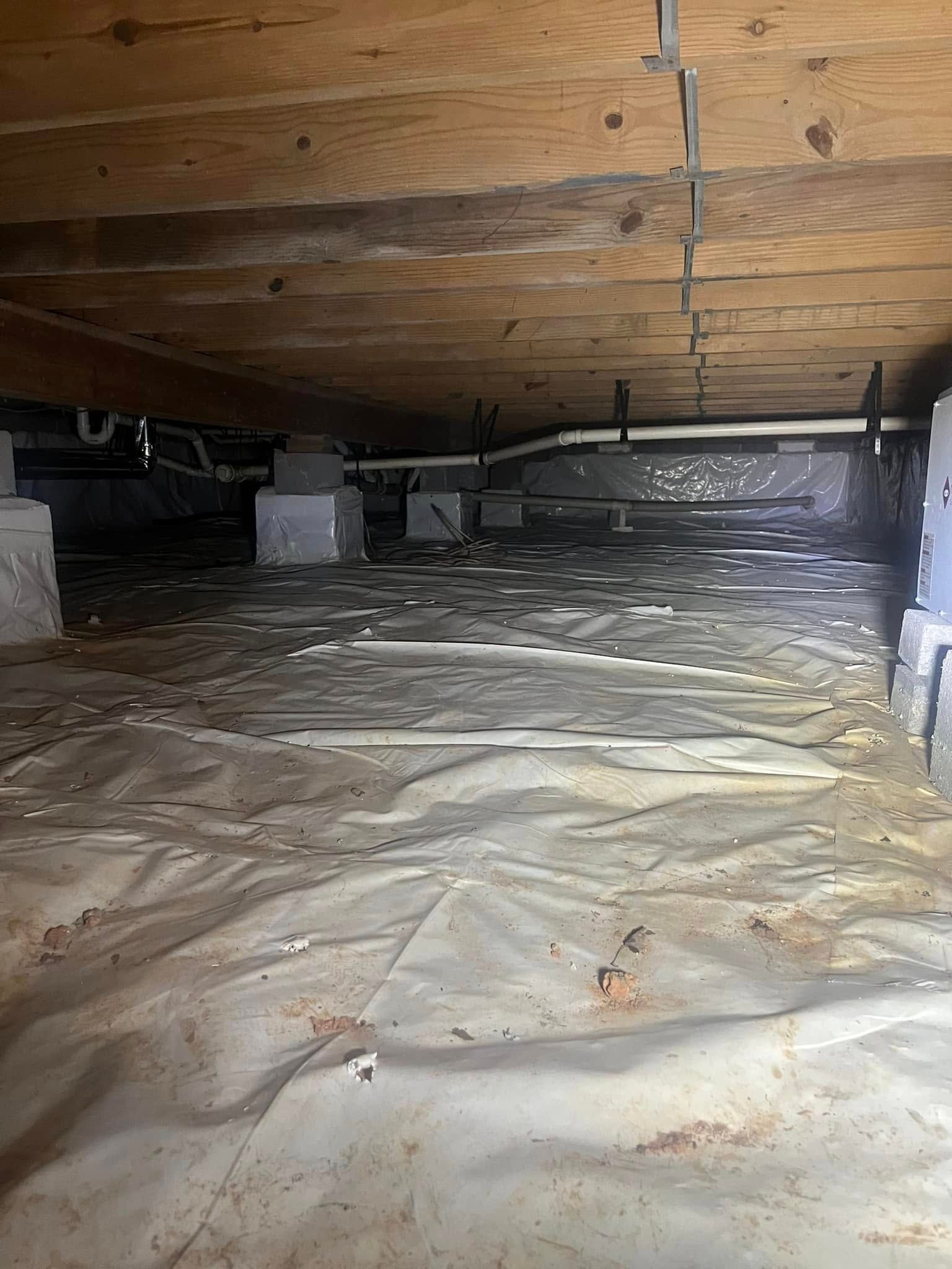 A low-angle view inside a crawl space featuring a white vapor barrier covering the ground, wooden joists, and pipes above.