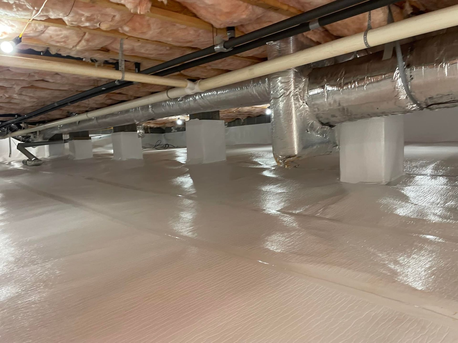 A low-angle view of a clean crawl space with a white vapor barrier, concrete support pillars, and overhead ductwork.