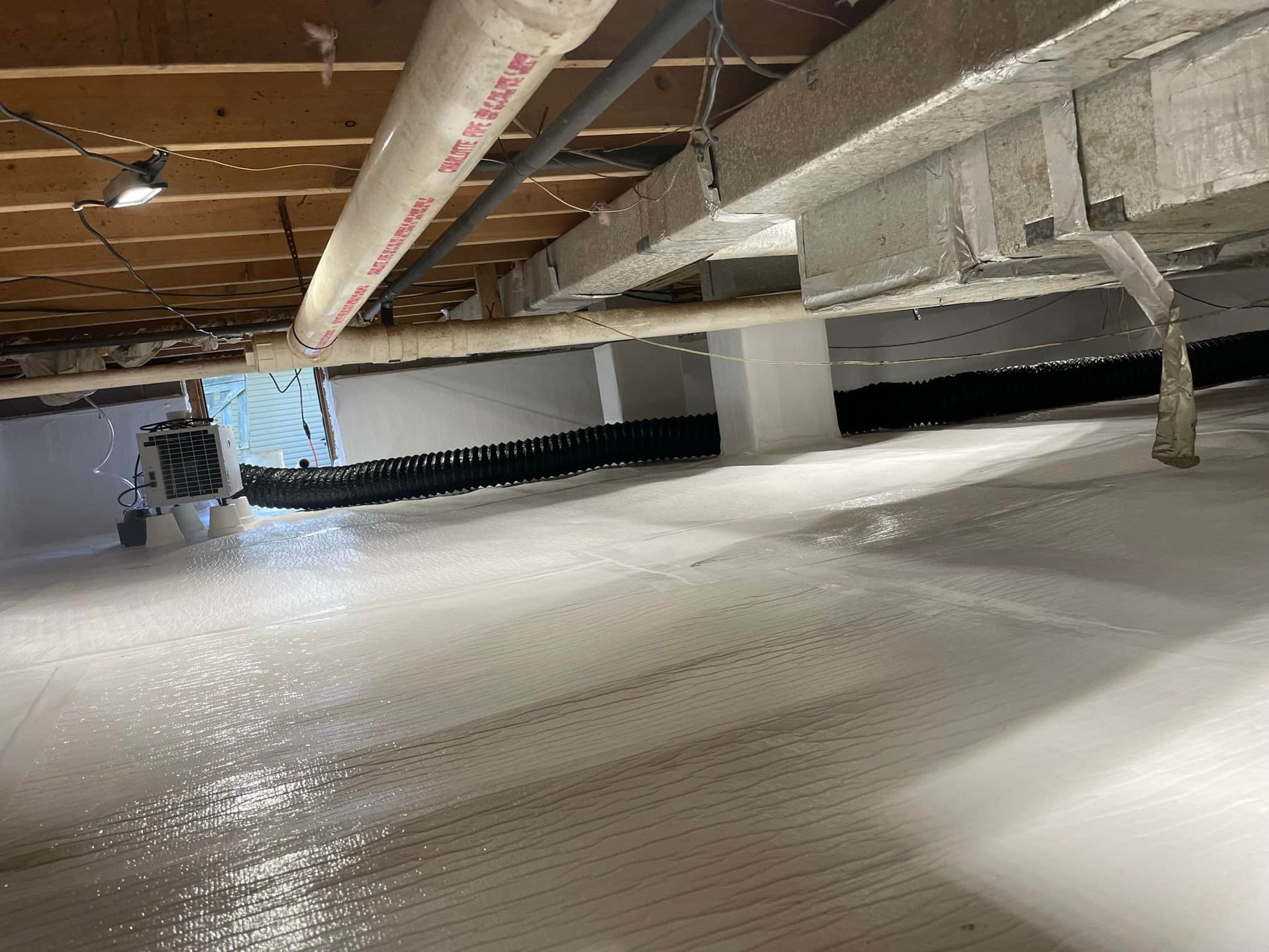 A clean, white-encapsulated crawl space with exposed wooden joists, pipes, a dehumidifier, and a black drainage pipe.