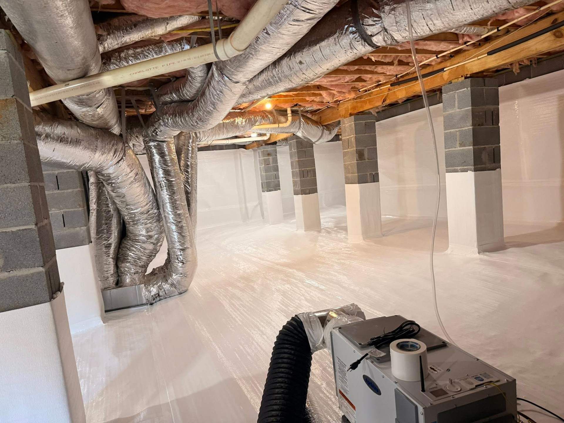 Crawl space with insulated silver ducts, white floor vapor barrier, and a dehumidifier unit in the foreground.