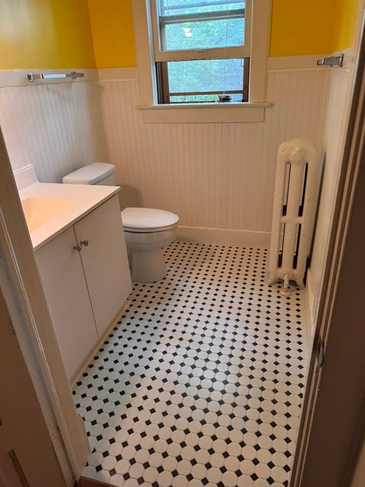 A bathroom with yellow walls, white beadboard wainscoting, a white vanity, a toilet, and a white radiator on tiled floors.