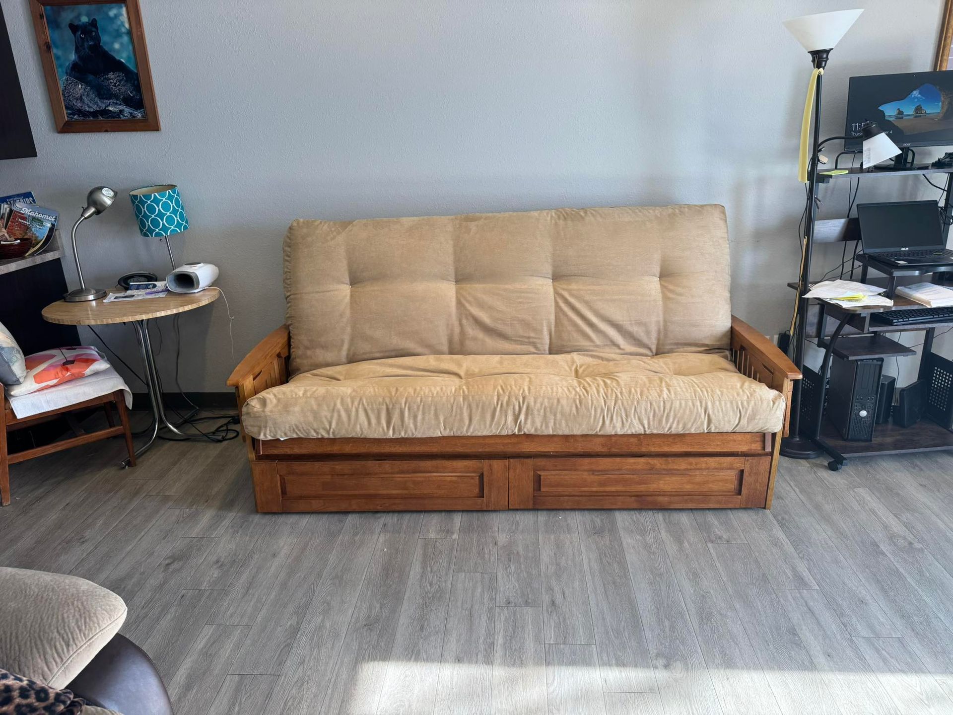 A beige futon with a wooden base centered in a room, flanked by a side table with a lamp and a computer desk.