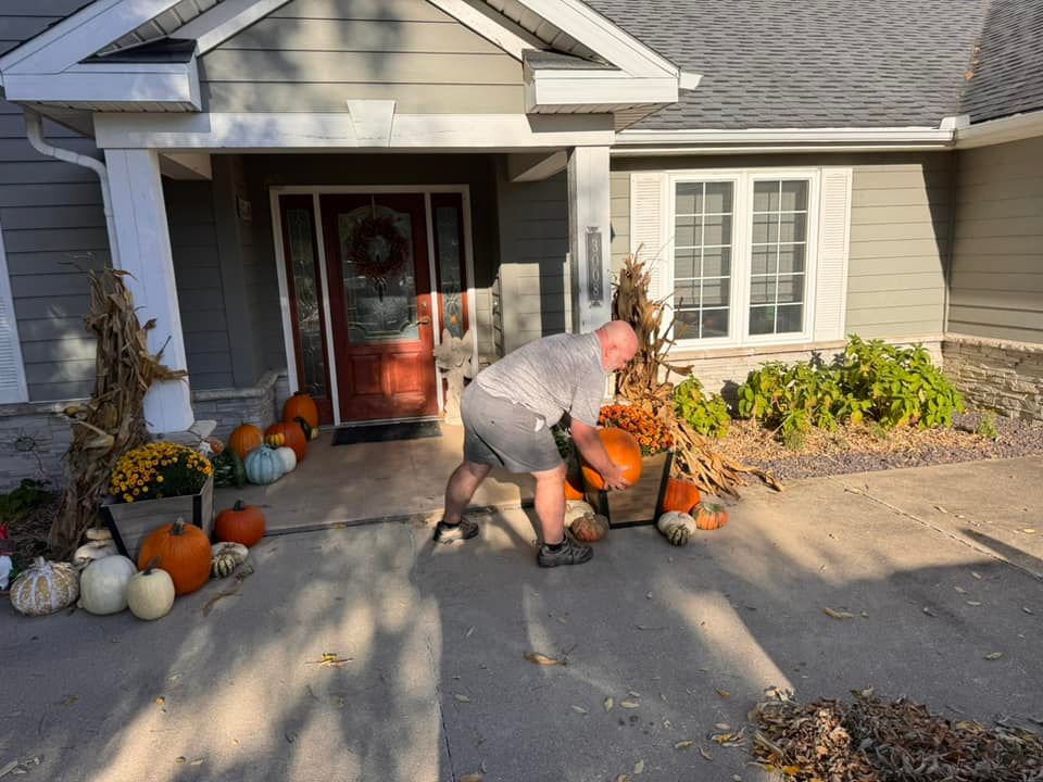 A person arranges pumpkins and fall decor in baskets near the front entrance of a house on a sunny day.