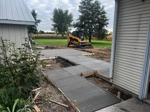 A yellow skid steer sits behind a newly poured concrete patio and walkway between two buildings in a grassy yard.