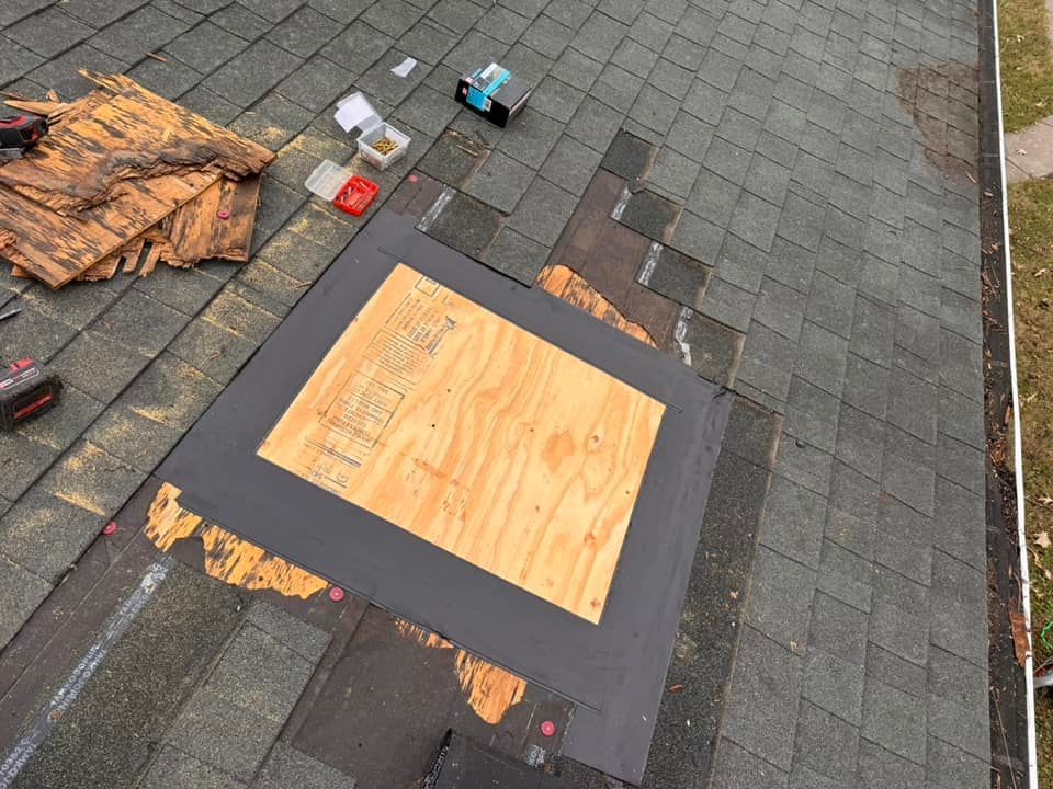 A rectangular patch of new plywood is installed on a shingled roof, surrounded by roofing underlayment and tools.