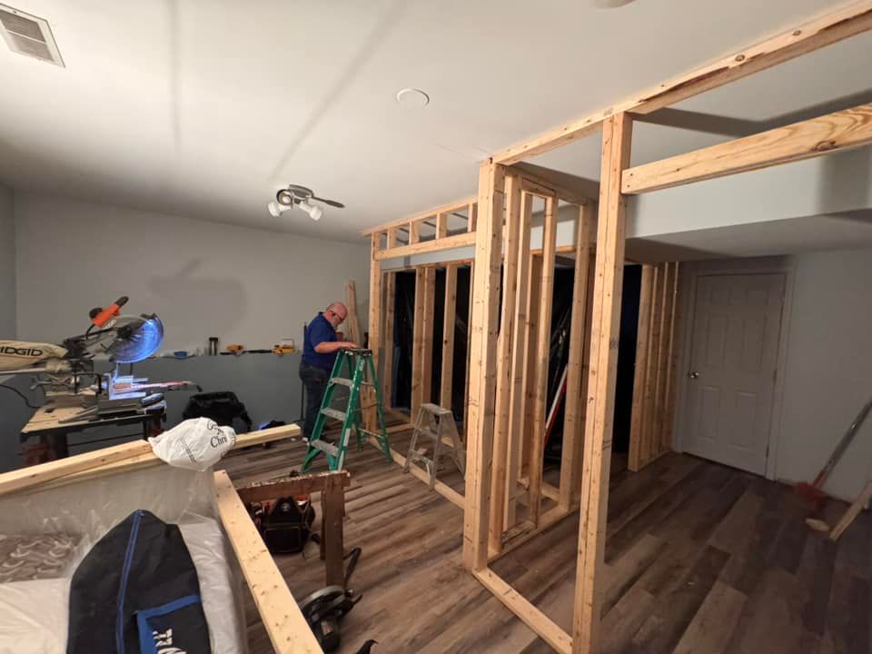 A person on a ladder works on wooden wall framing in a room with light-colored walls and wood-patterned flooring.