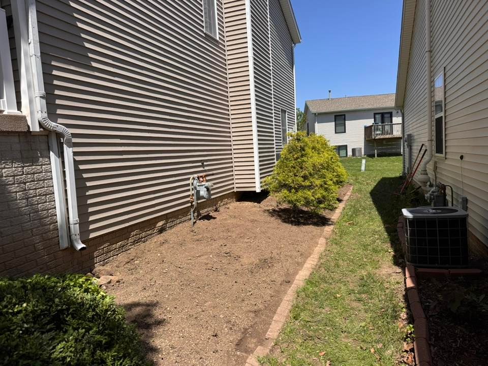 A side yard between two beige vinyl-sided houses, featuring a dirt patch, a small yellow-green bush, and an AC unit.