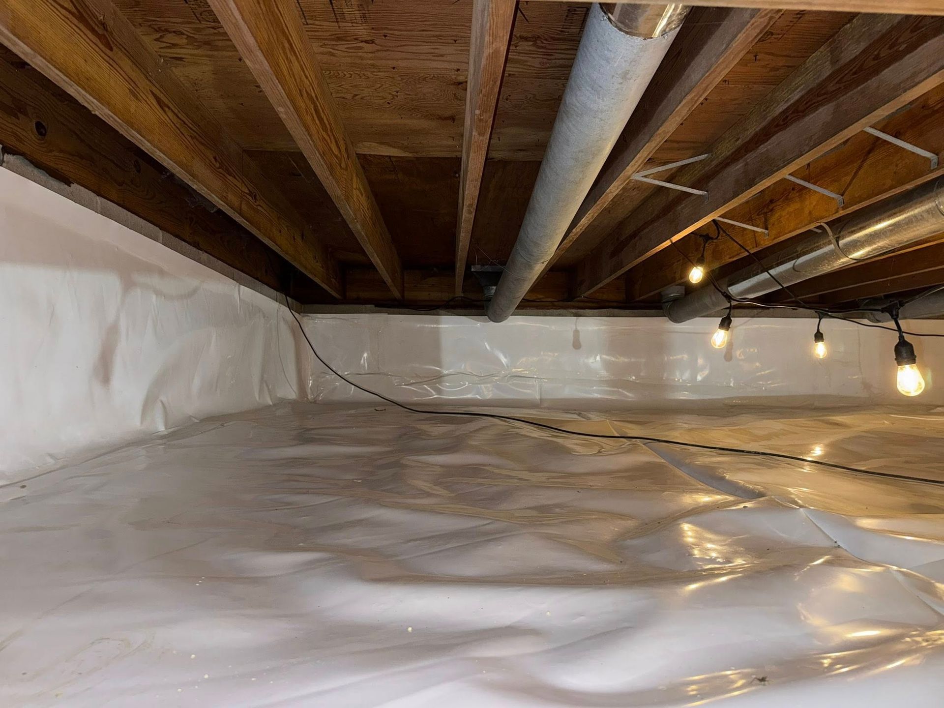 A crawl space with white vapor barrier lining the floor and walls, exposed wooden floor joists, and hanging light bulbs.