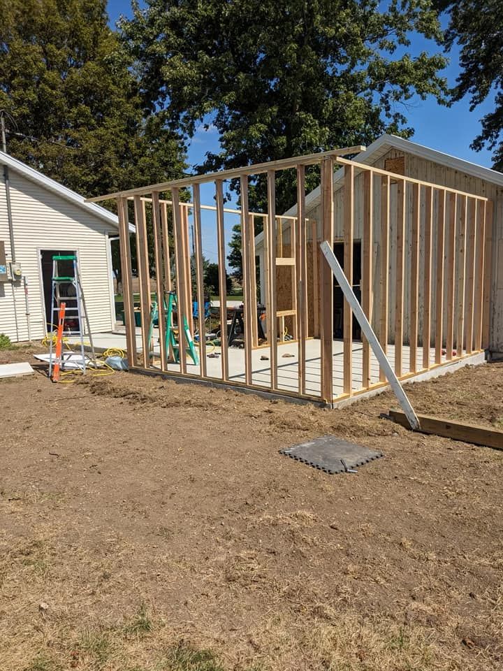 Wooden wall framing for a room addition under construction, attached to the side of an existing building on a sunny day.