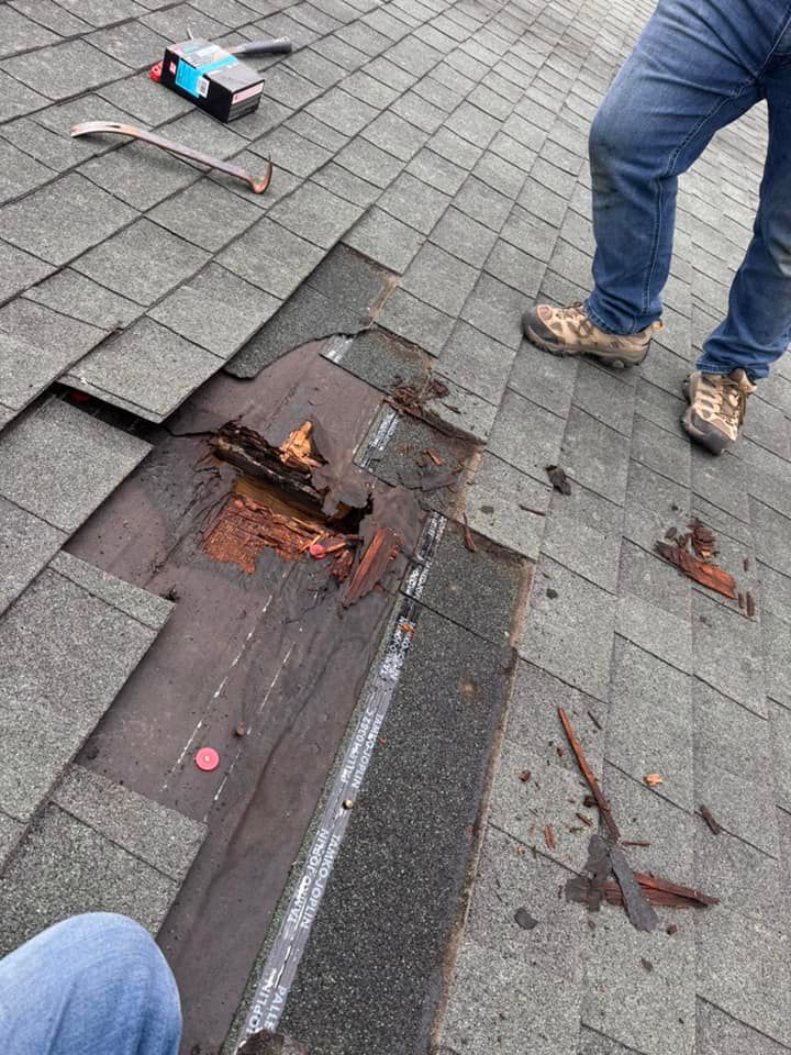 A person repairs a roof, showing rotted plywood deck exposed beneath removed asphalt shingles near tools and debris.